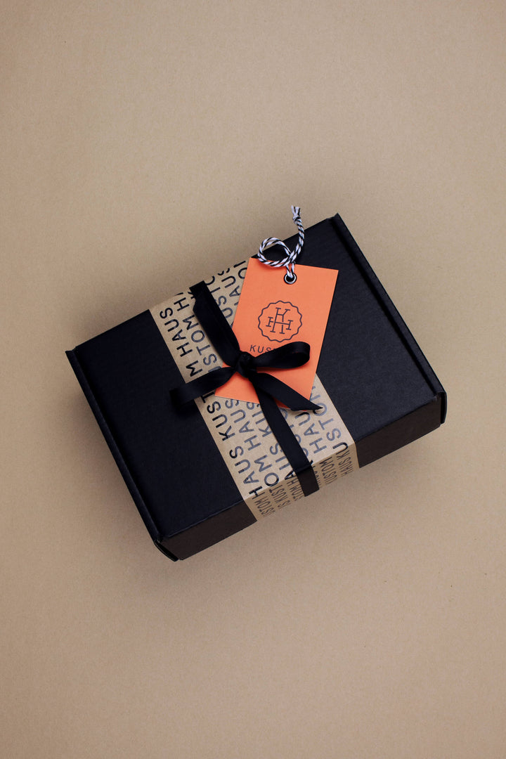A black rectangular gift box wrapped with a beige paper band printed with black text, tied with a black ribbon and decorated with an orange paper tag, on a solid beige background.