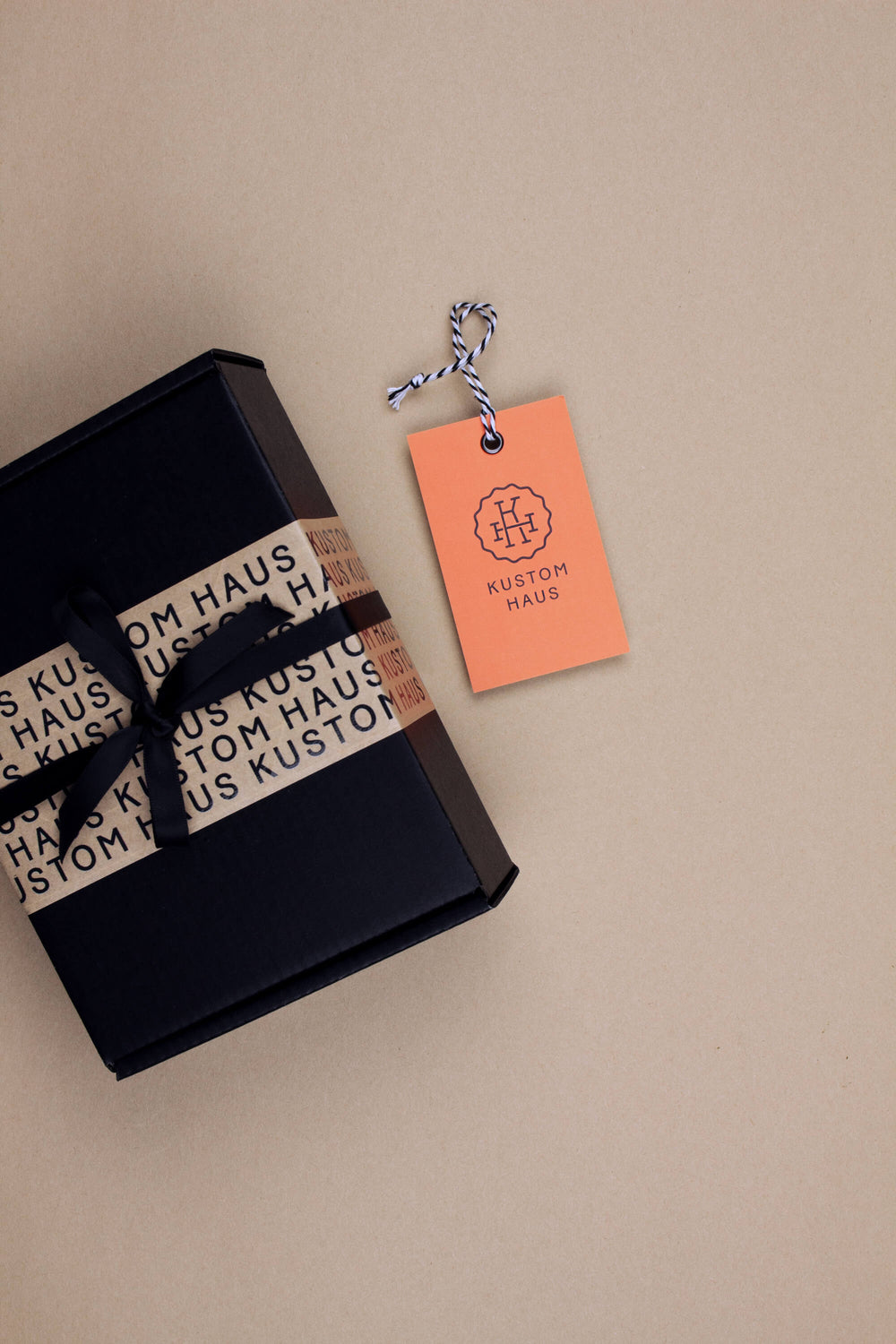 A black gift box wrapped with a black ribbon printed with the brand name 'KUSTOM HAUS' in black on a cream paper band, alongside a peach-colored paper hangtag with the same logo, on a solid beige background.