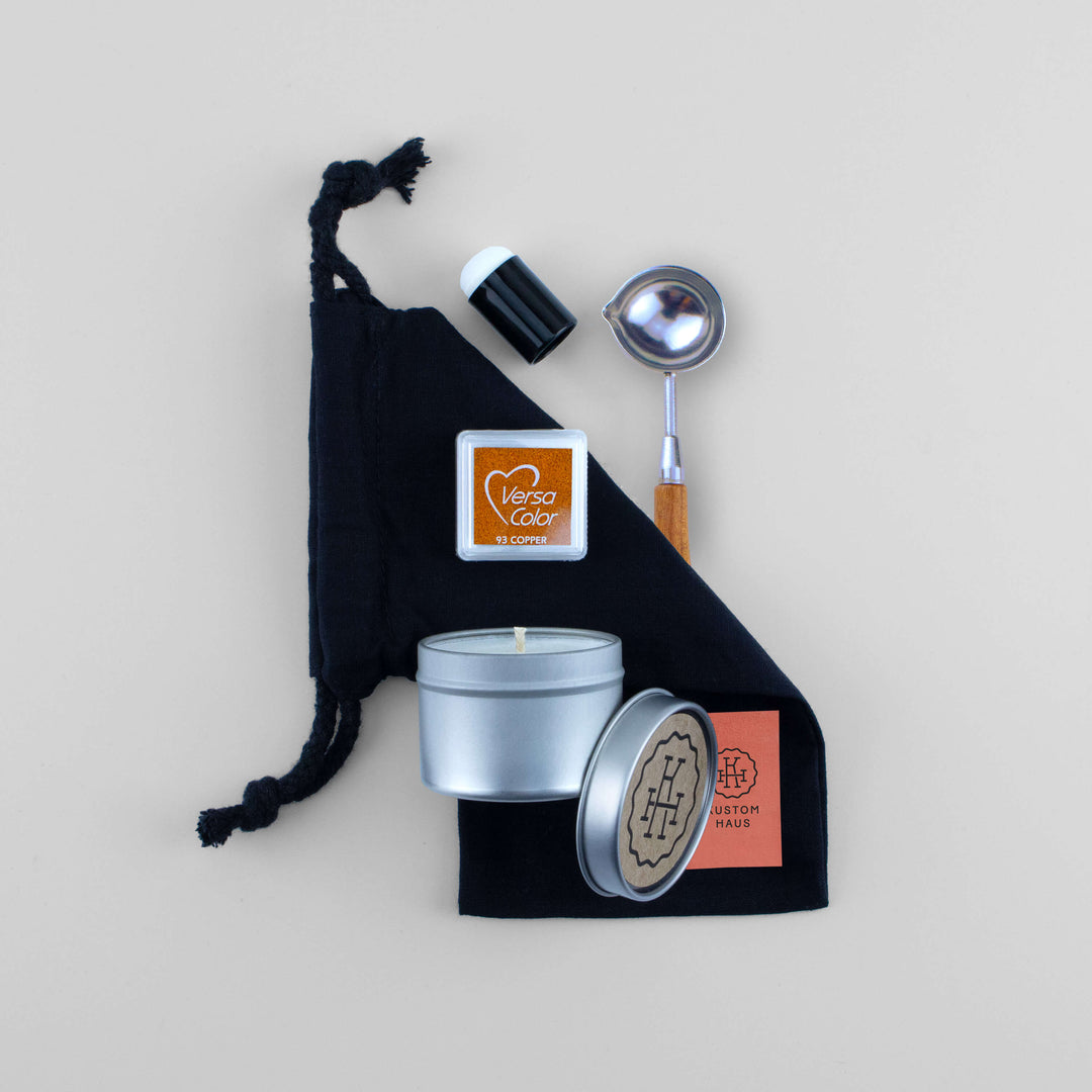 A wax melting kit gift set arranged on a light beige background, featuring a black drawstring bag, a silver tin candle, a small copper Versa Color highlighting ink pad, and a silver-and-wood spoon. The silver tin and lid show the brand Kustom Haus.