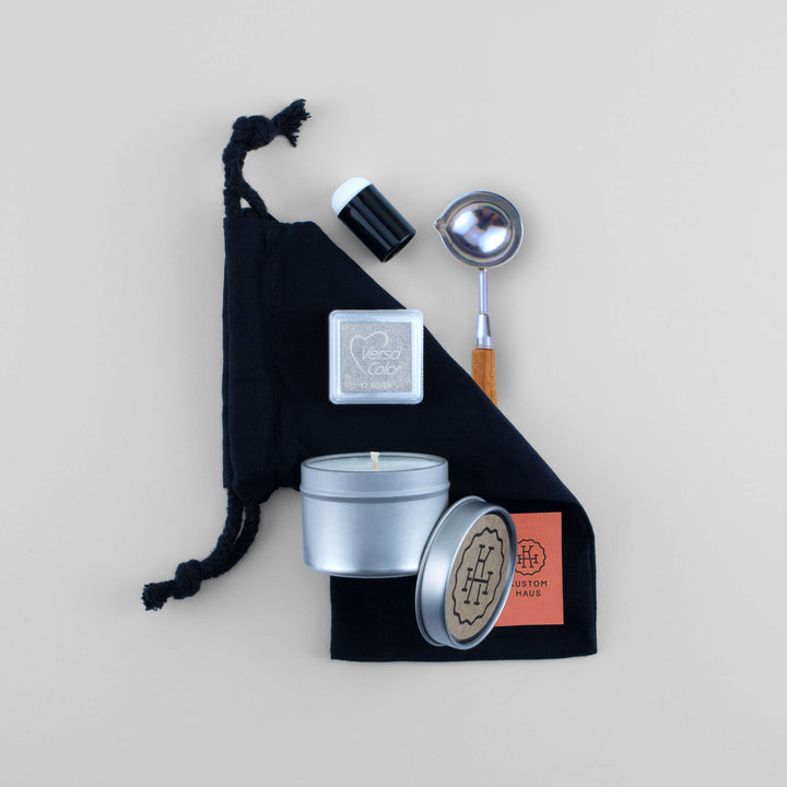 A wax melting kit gift set arranged on a light beige background, featuring a black drawstring bag, a silver tin candle, a small silver Versa Color highlighting ink pad, and a silver-and-wood spoon. The silver tin and lid show the brand Kustom Haus.
