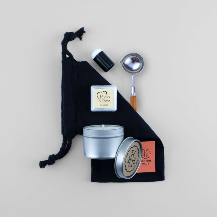 A wax melting kit gift set arranged on a light beige background, featuring a black drawstring bag, a silver tin candle, a small white Versa Color highlighting ink pad, and a silver-and-wood spoon. The silver tin and lid show the brand Kustom Haus.